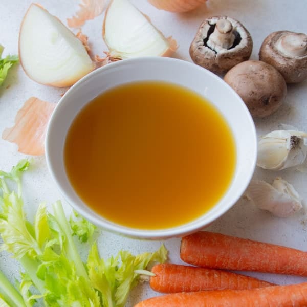 vegetable broth in a white bowl surrounded by carrots, onions, mushrooms, and celery
