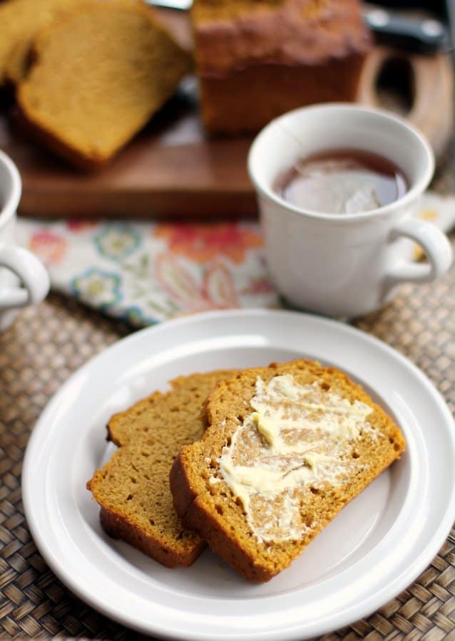 slices of pumpkin cider bread with butter on a white plate next to a cup of tea