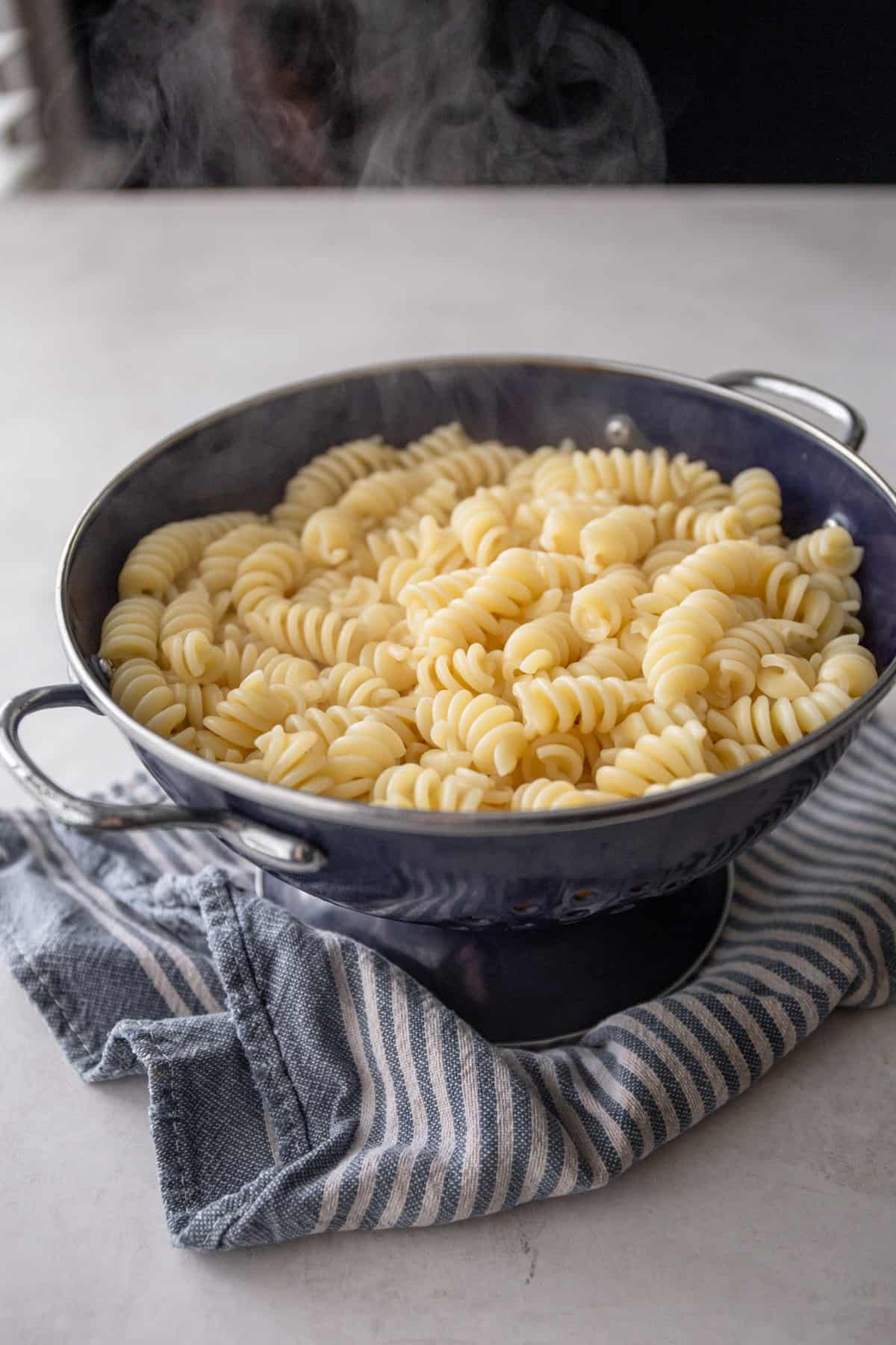 cooked pasta in a blue strainer on a blue and grey stripped dish towel