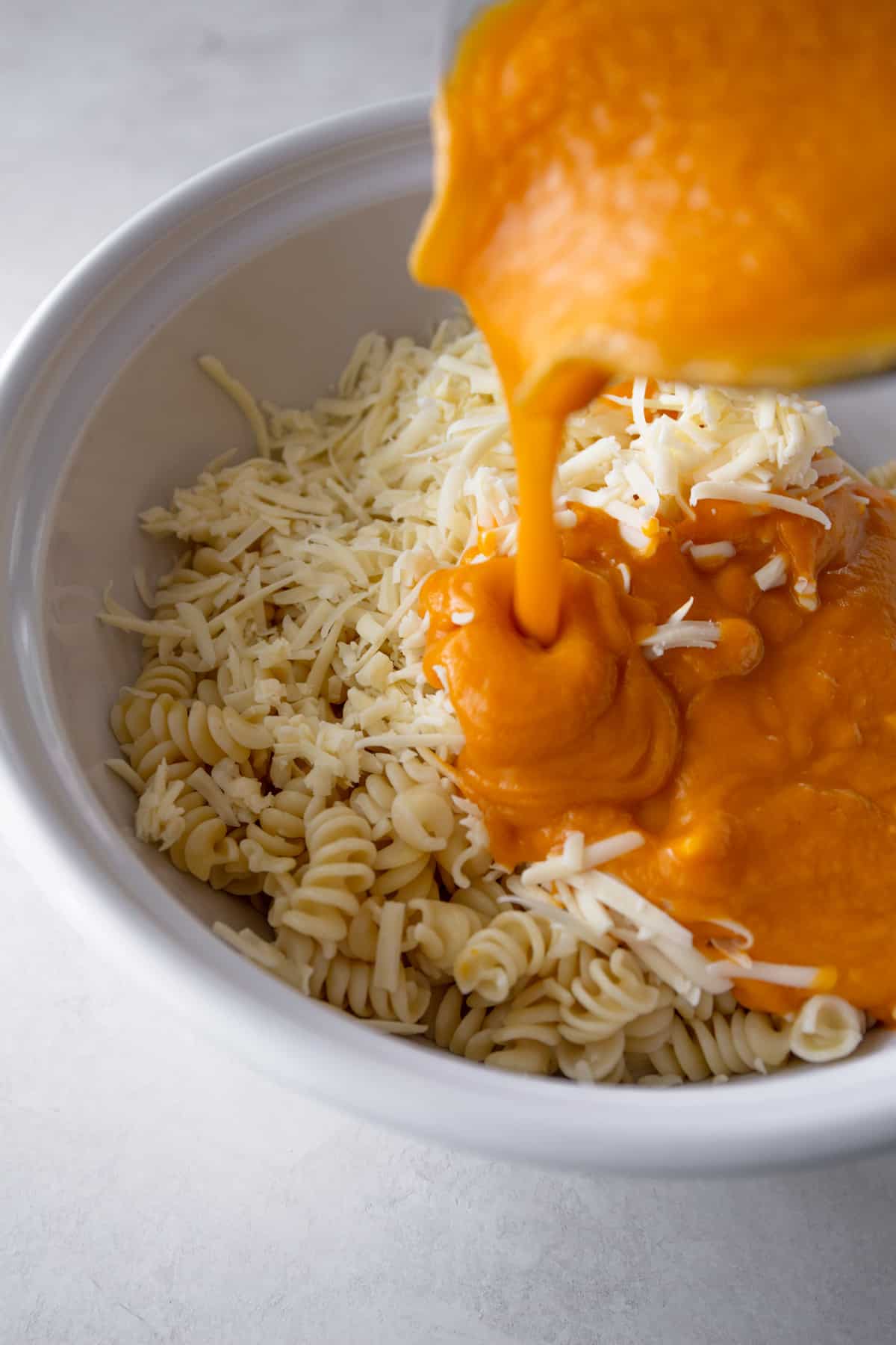 Pouring sweet potato puree over pasta and cheese in a large white mixing bowl