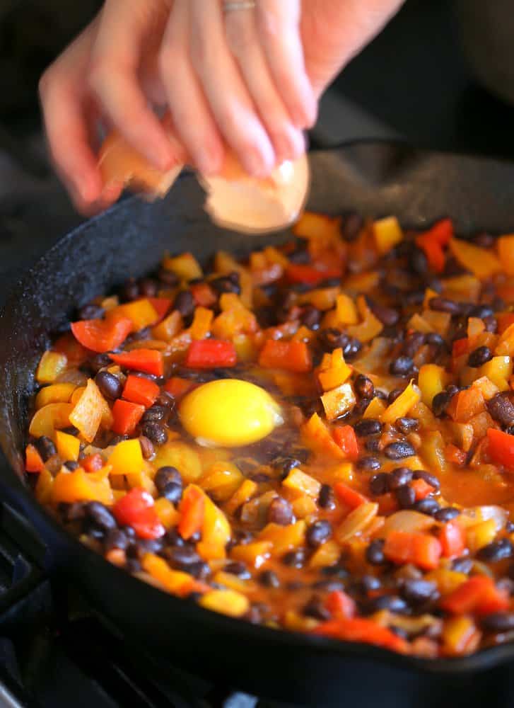 cracking eggs over mixed vegetables in a cast iron skillet