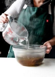a Caucasian person in a green apron pouring hot water over tamarind pulp in a glass bowl