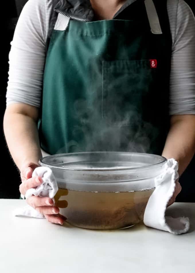 a person in a dark green apron holding a steaming glass bowl of soaking tamarind pulp