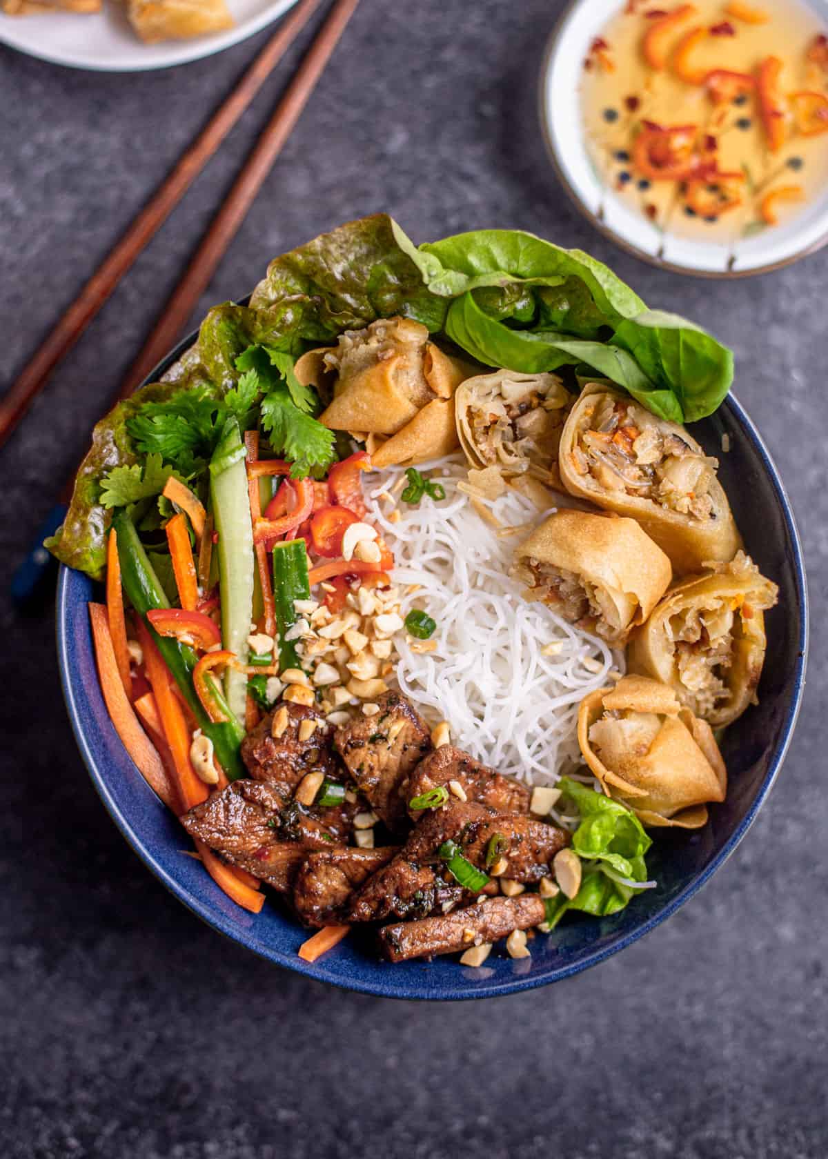 overhead image of rice noodles in blue bowl topped with lemongrass pork, vegetables, and spring rolls