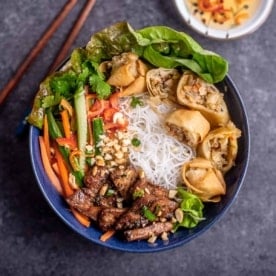 square image of overhead image of rice noodles in blue bowl topped with lemongrass pork, vegetables, and spring rolls