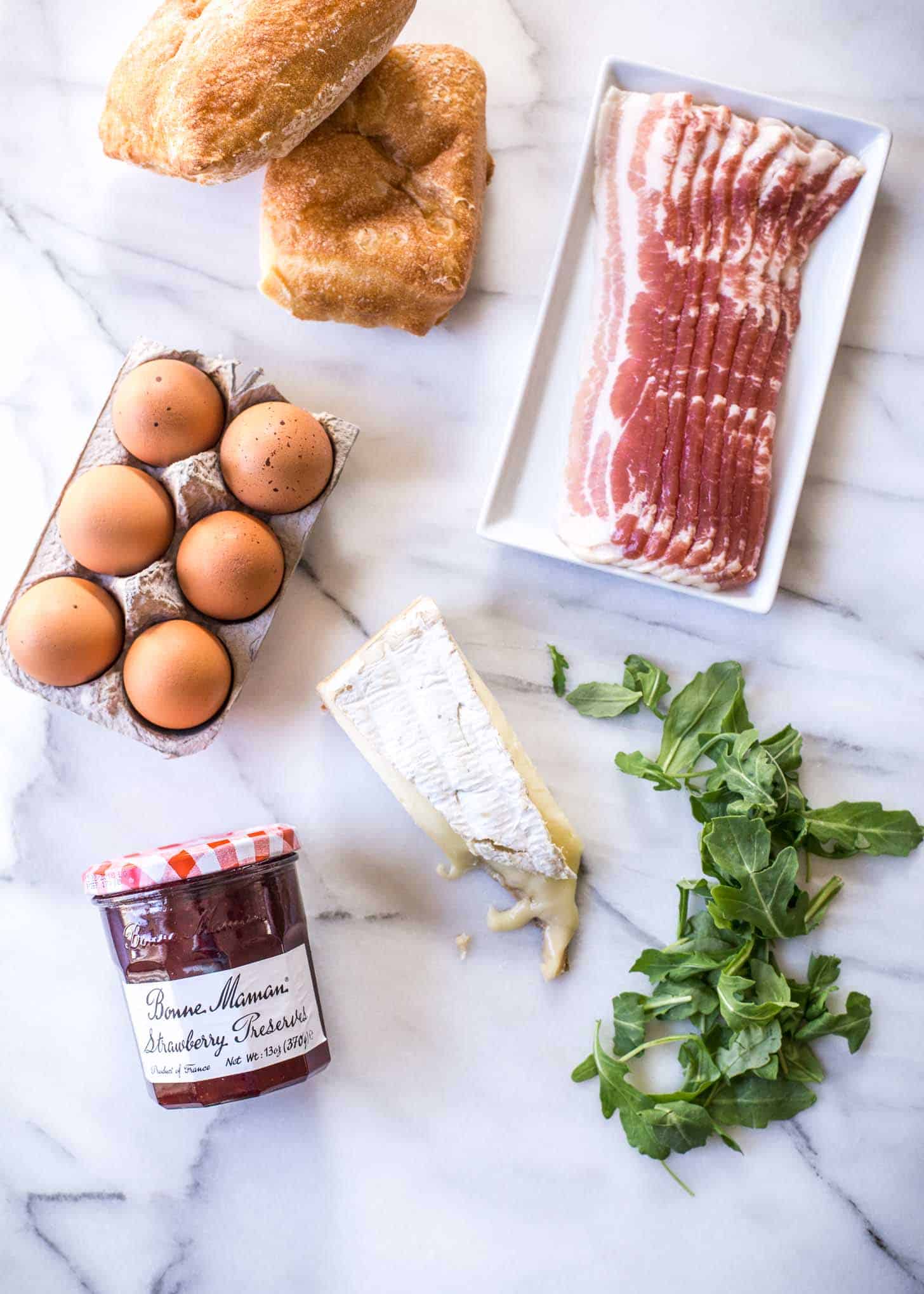 bread, bacon, eggs, brie and strawberry preserves on a white countertop