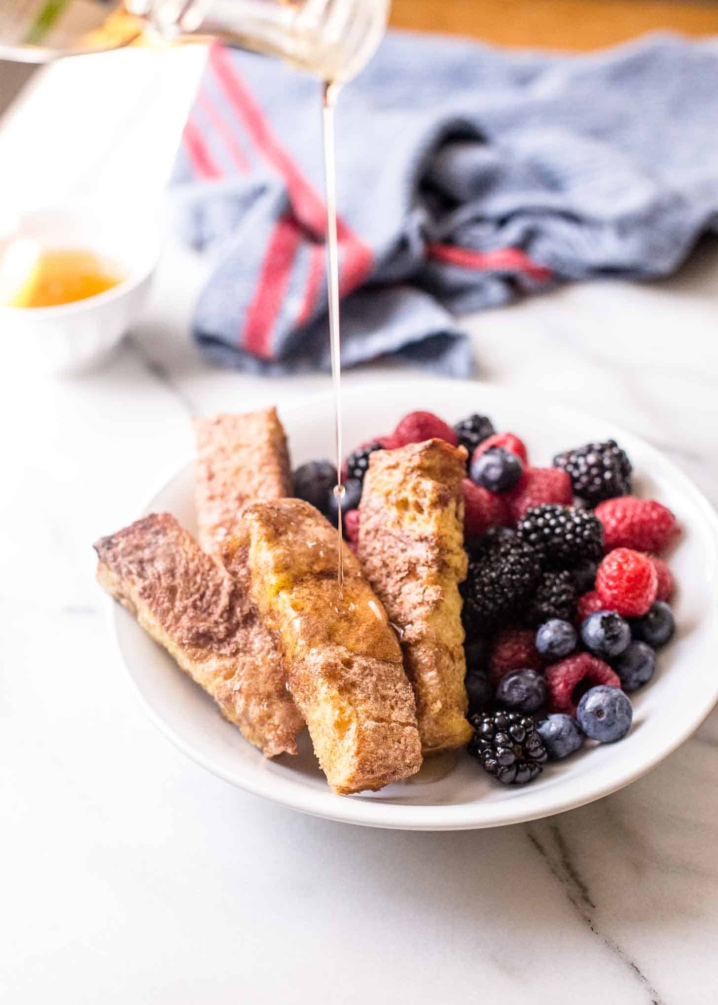 drizzling syrup on french toast sticks with berries in a white bowl