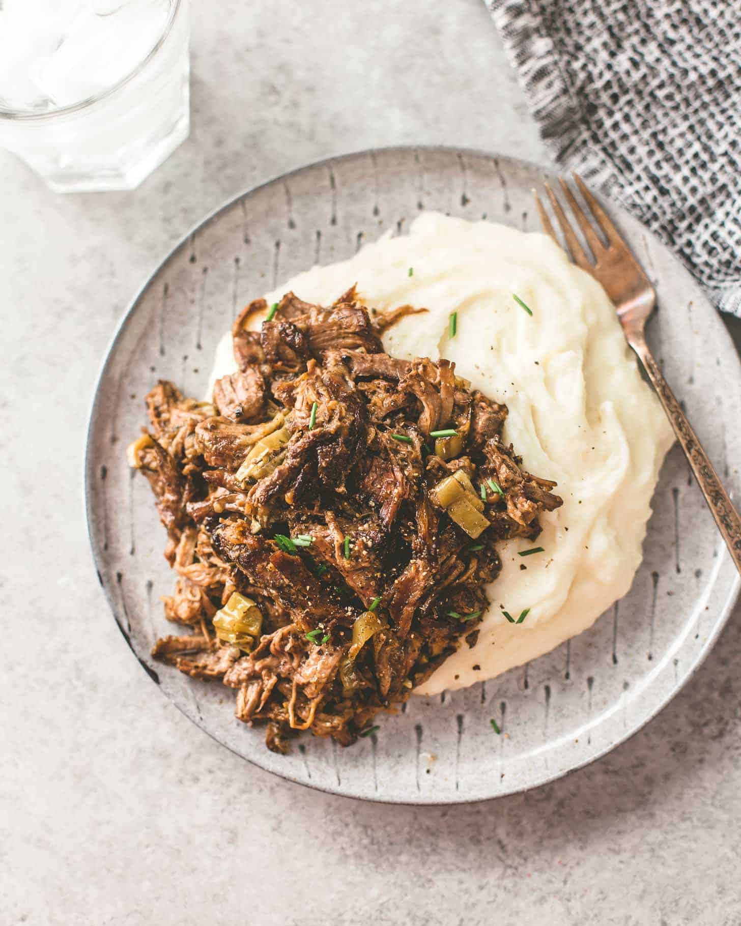 overhead image of Slow Cooker Mississippi Roast over mashed potatoes on a grey plate with a fork