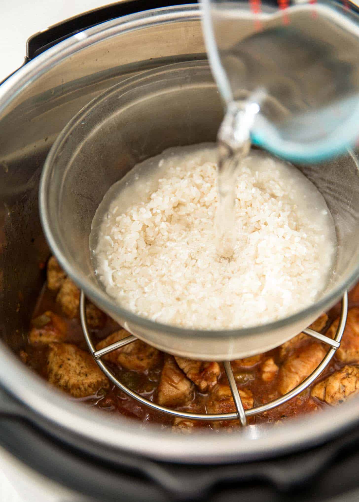 overhead image of chicken cooking in an Instant Pot with a rack and a clear glass bowl. There is a glass measuring cup pouring water over the rice in the bowl. 