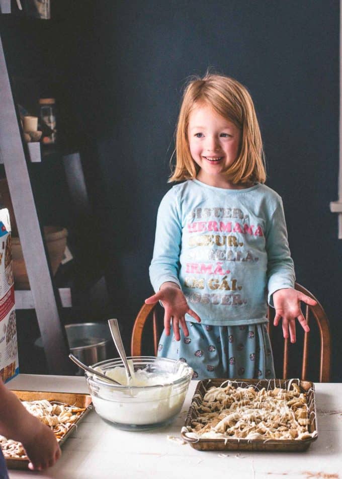 a young girl with a sheet pan of easter egg crunch bark on a white table, next to a bowl of melted almond bark
