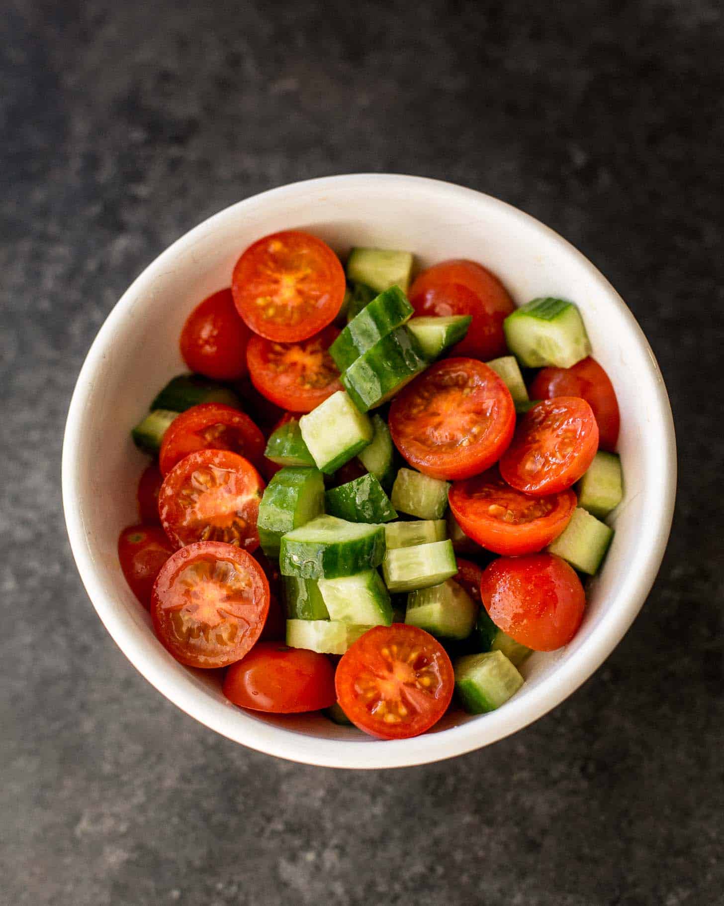 overhead image of cut up cucumbers and tomatoes in a white bowl on a grey countertop