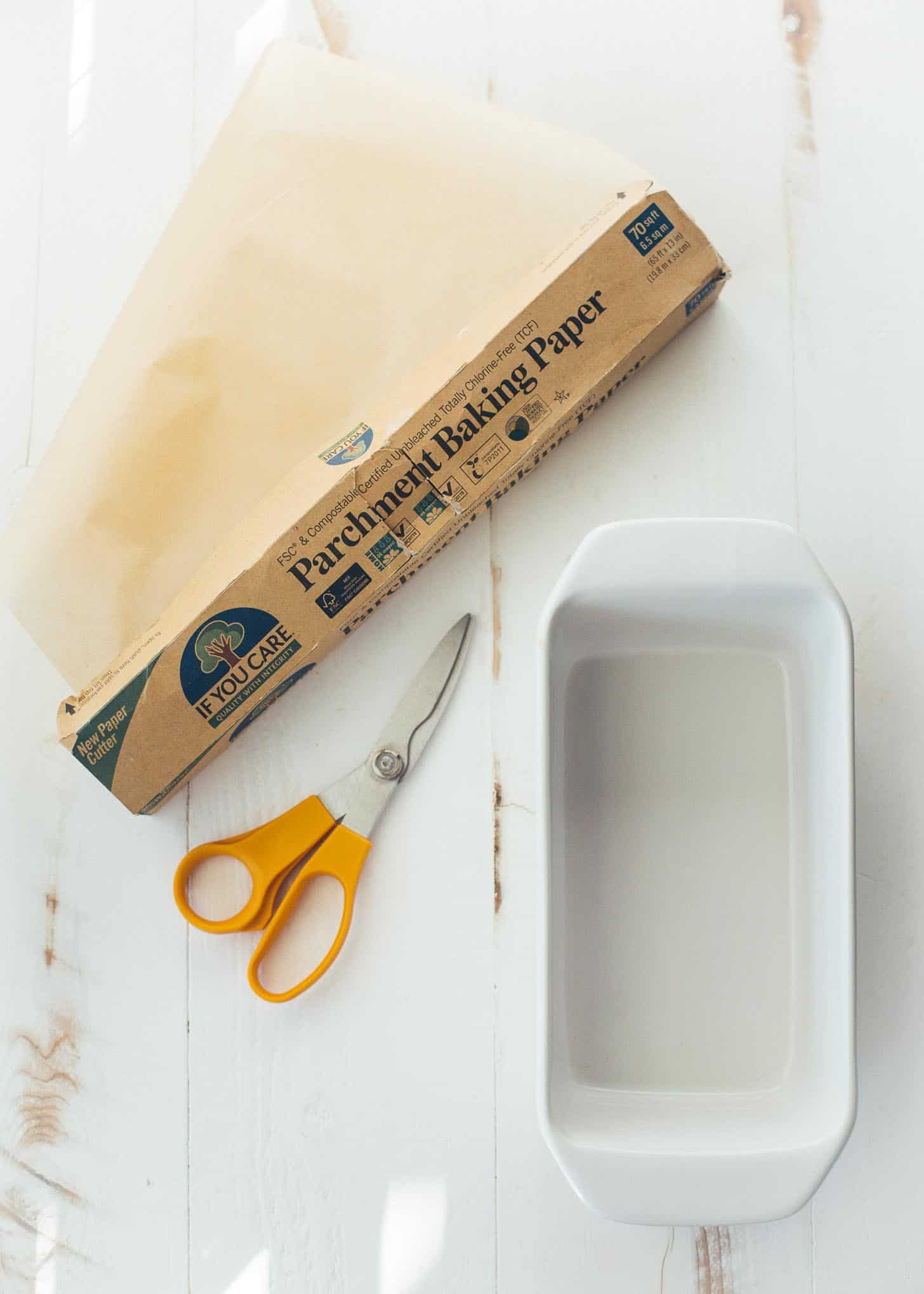 overhead image of a roll of parchment paper and scissors on a white table next to a loaf pan