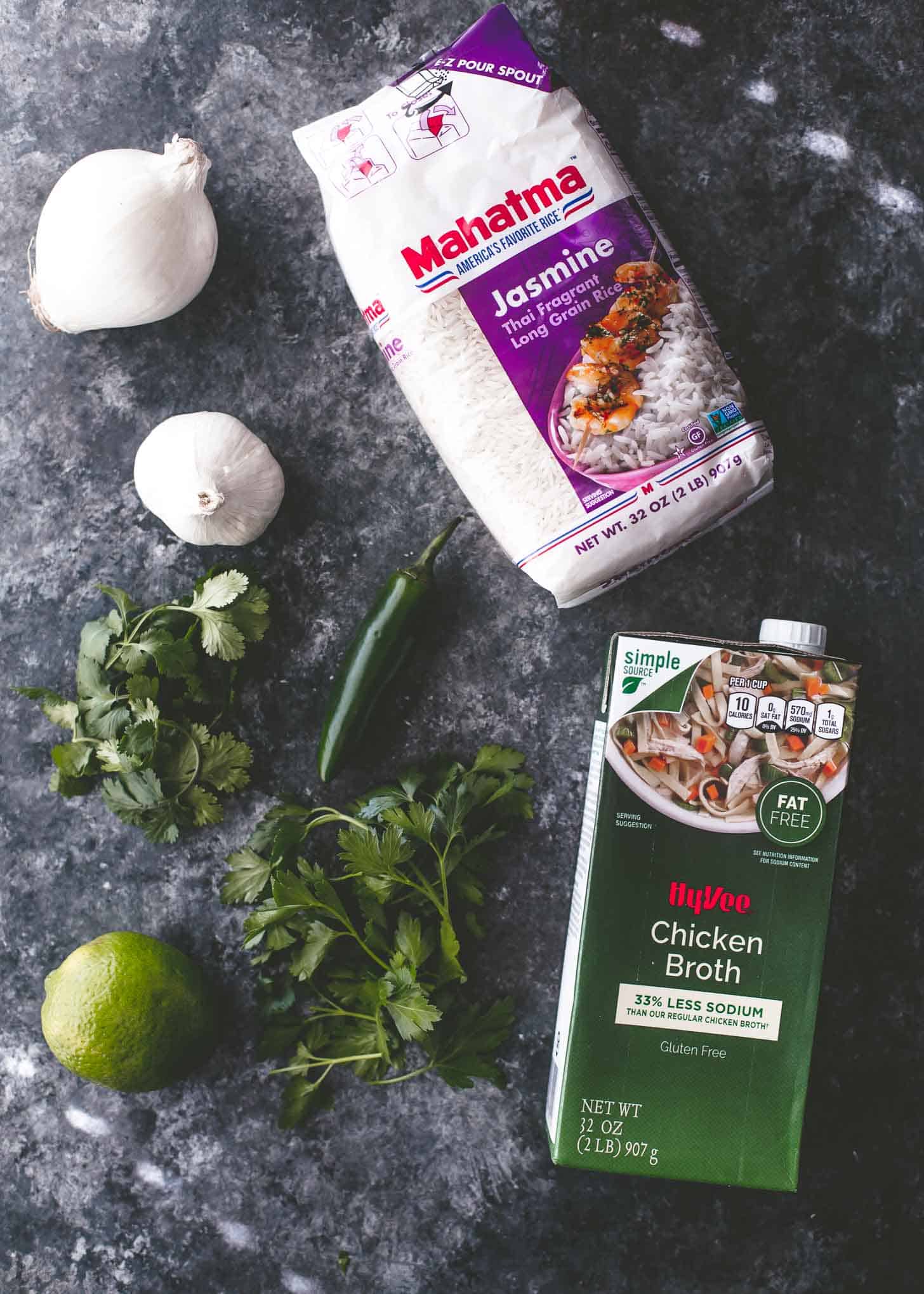 overhead image of jasmine rice, chicken broth, garlic, jalapeno, lime and herbs on a grey table