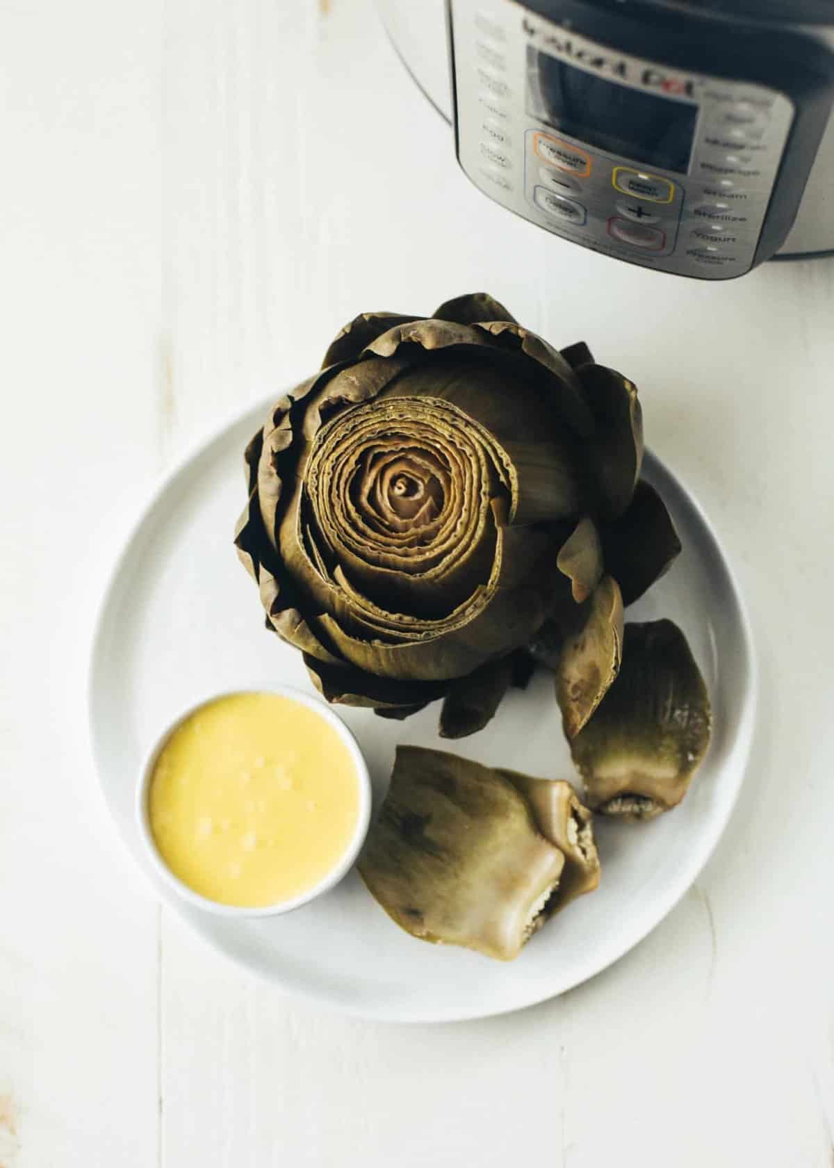 Instant Pot Steamed Artichoke on a white plate next to a bowl of hollandaise