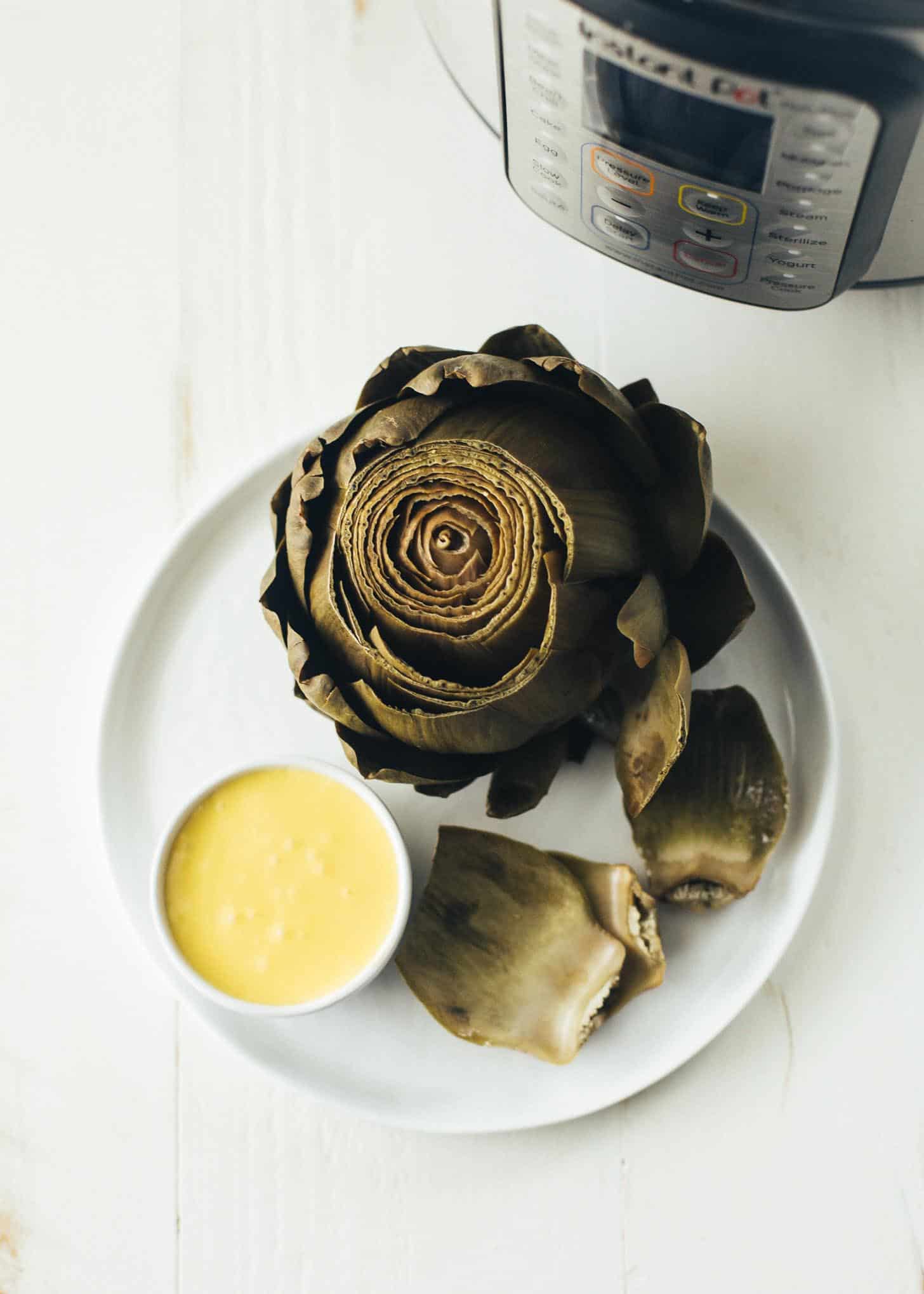 overhead image of Instant Pot Steamed Artichoke on a white plate, next to a small white bowl of hollandaise sauce