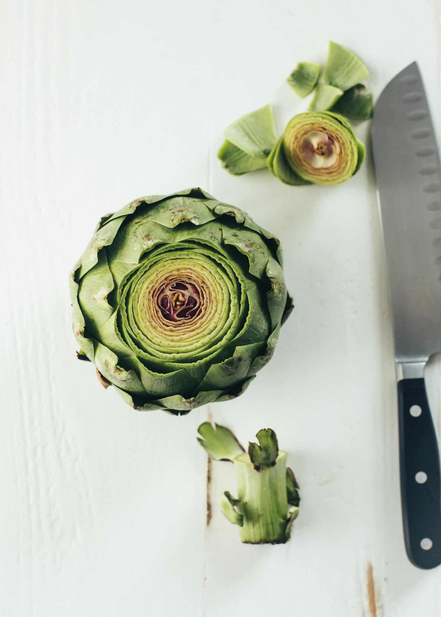 an artichoke with the top sliced off next to a knife on a white table