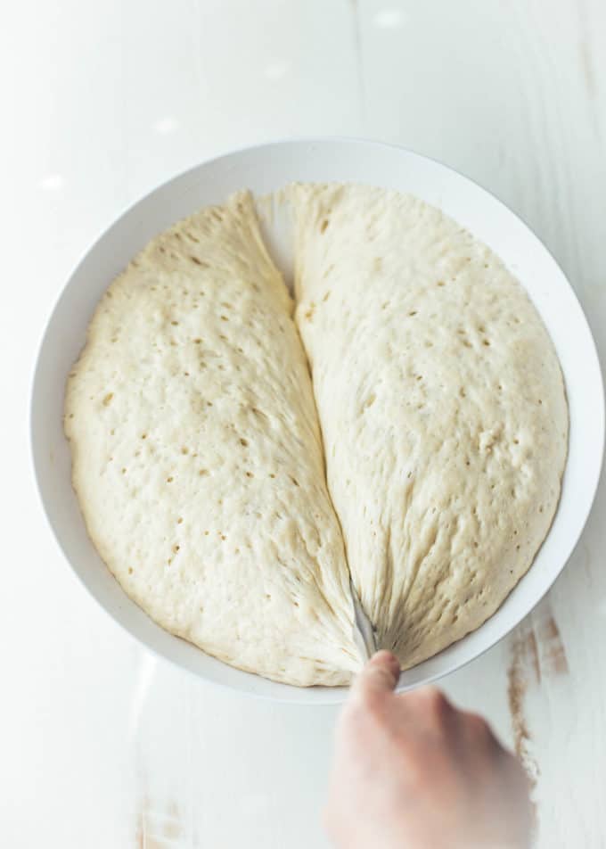 slicing risen focaccia dough in half in a white bowl