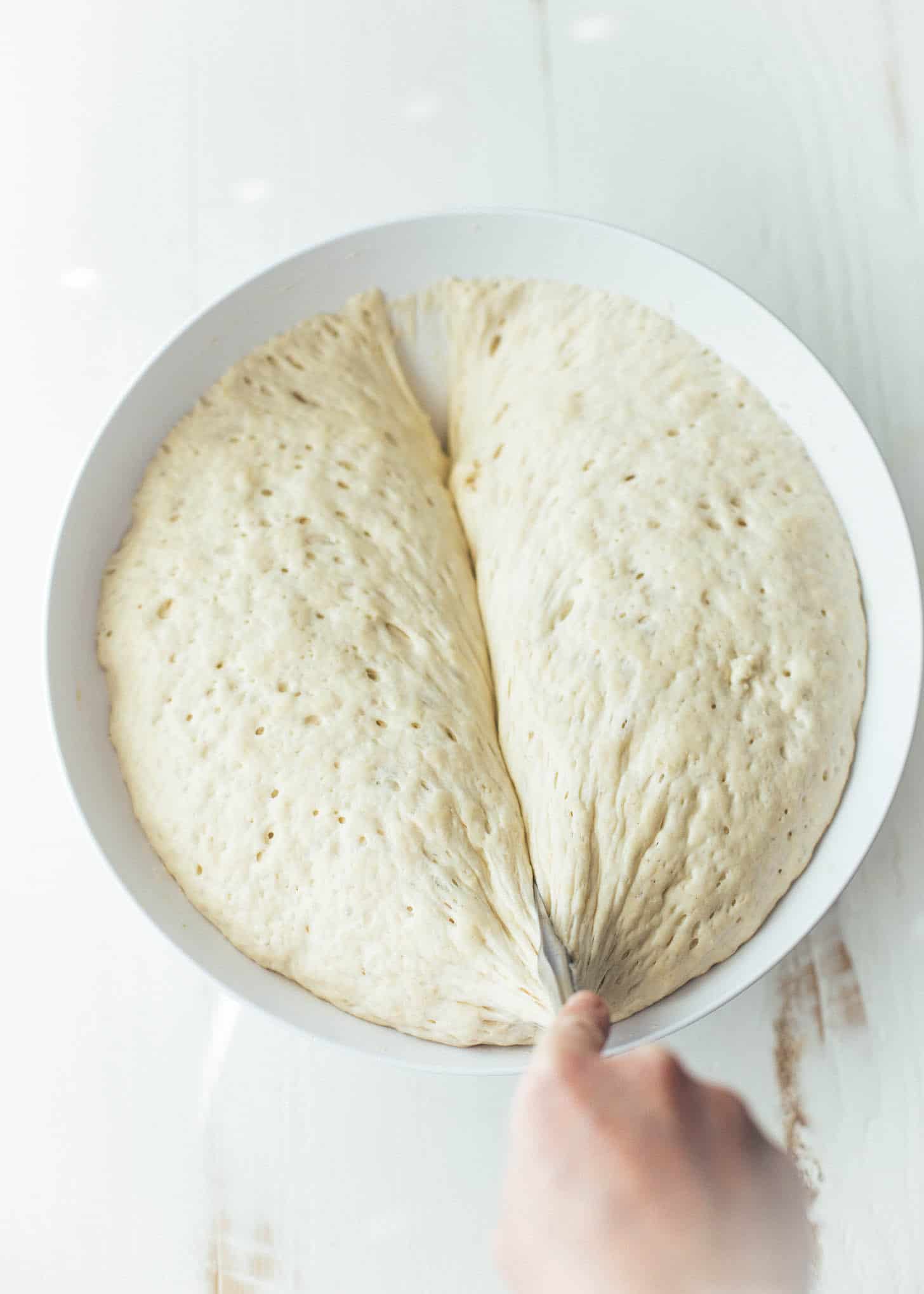 overhead image of slicing risen focaccia dough in half in a white bowl