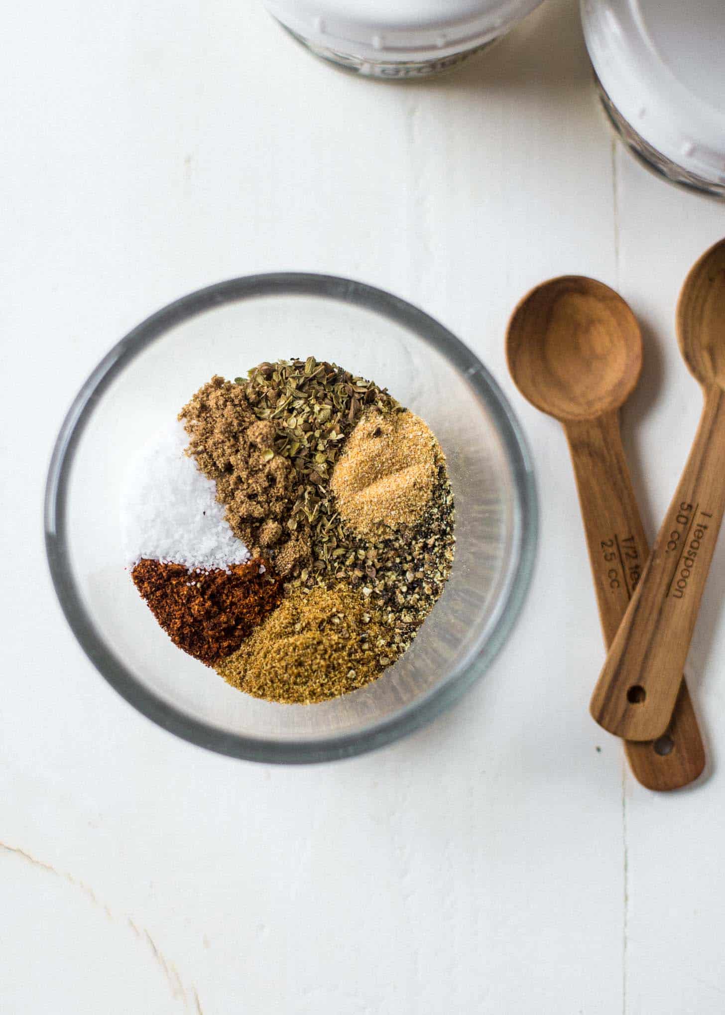 overhead image of dried spices in a clear bowl, next to wooden measuring spoons on a white table