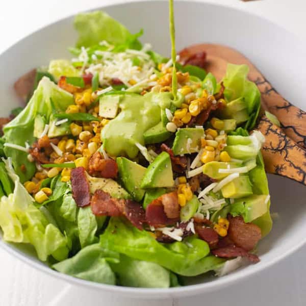 a white bowl with lettuce, bacon, avocado, corn and a green herb vinaigrette being poured over top