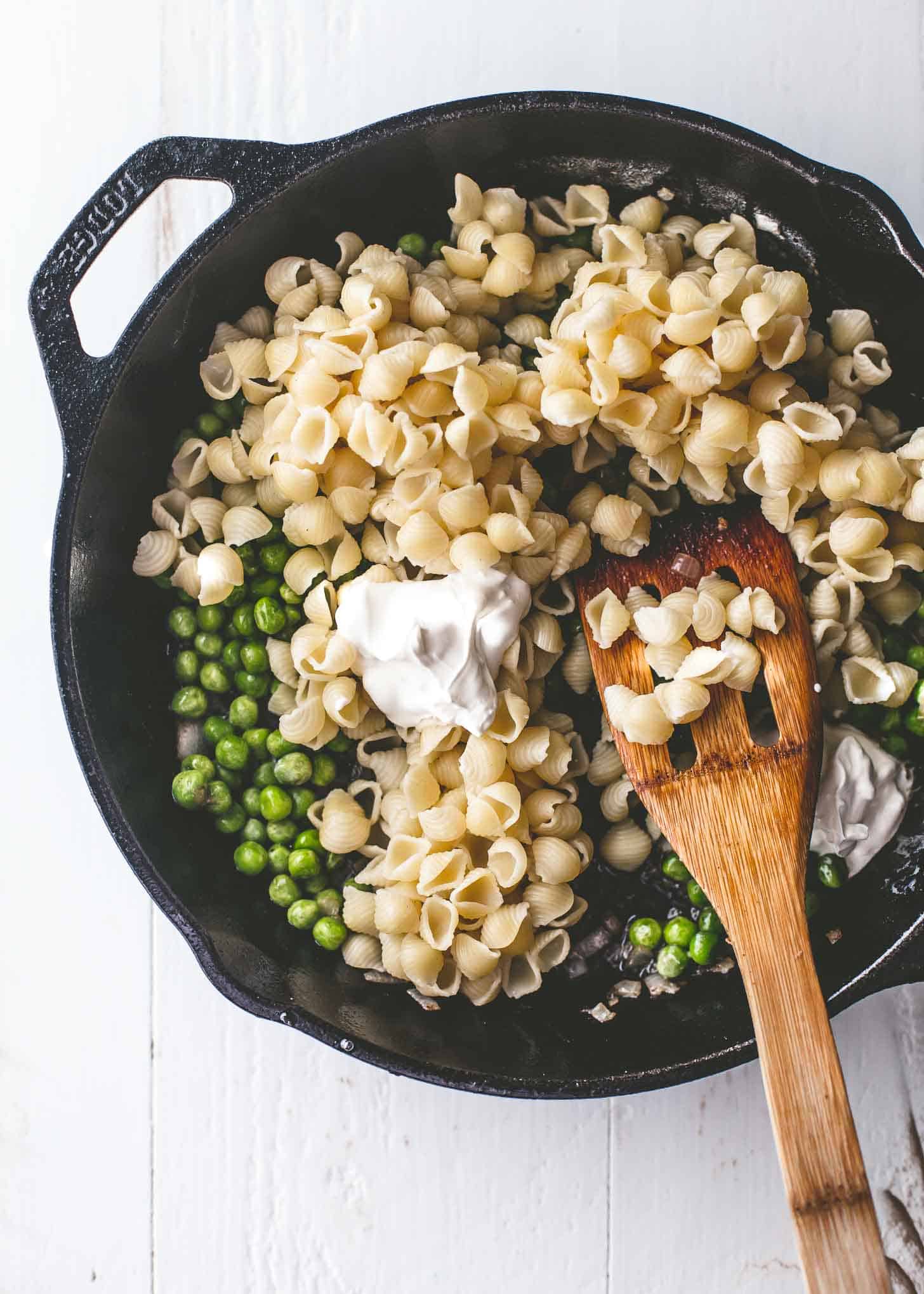 overhead image of mixing shells, peas and creme fraiche in a cast iron skillet with a wooden spoon