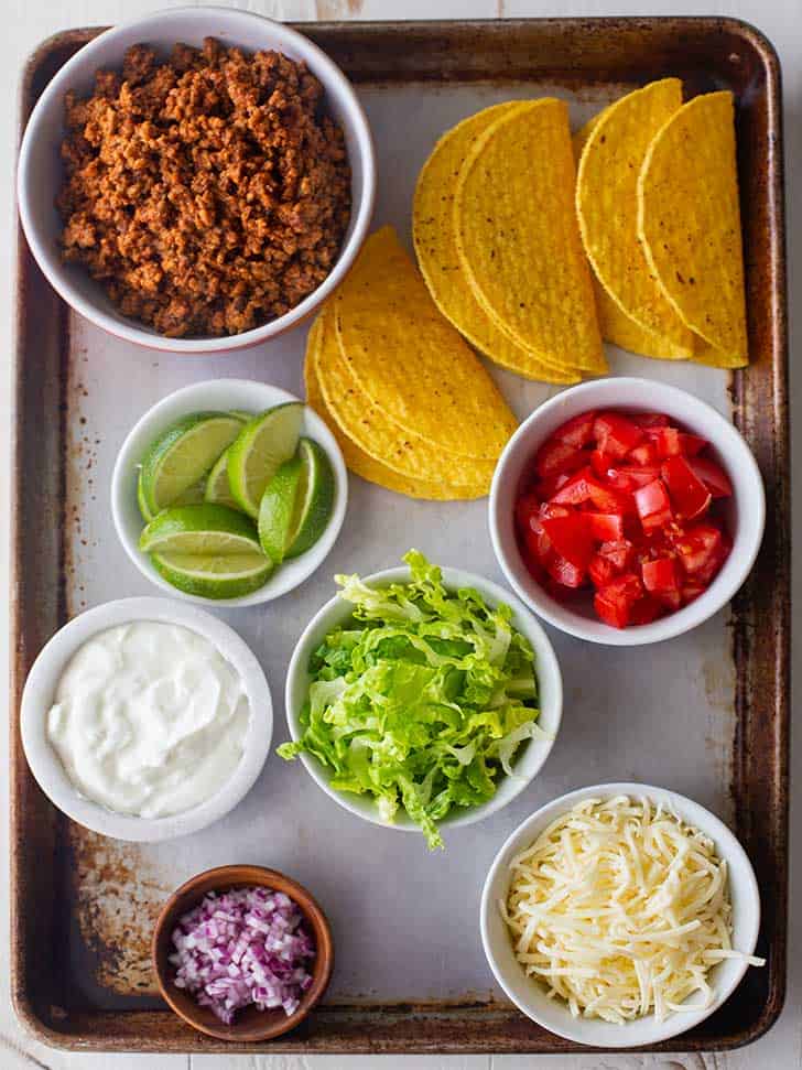 taco shells, ground beef, limes, lettuce, tomatoes, sour cream, cheese, and onions on a tray