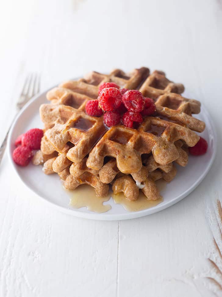 Overnight whole wheat waffles on a white plate, topped with raspberries and syrup
