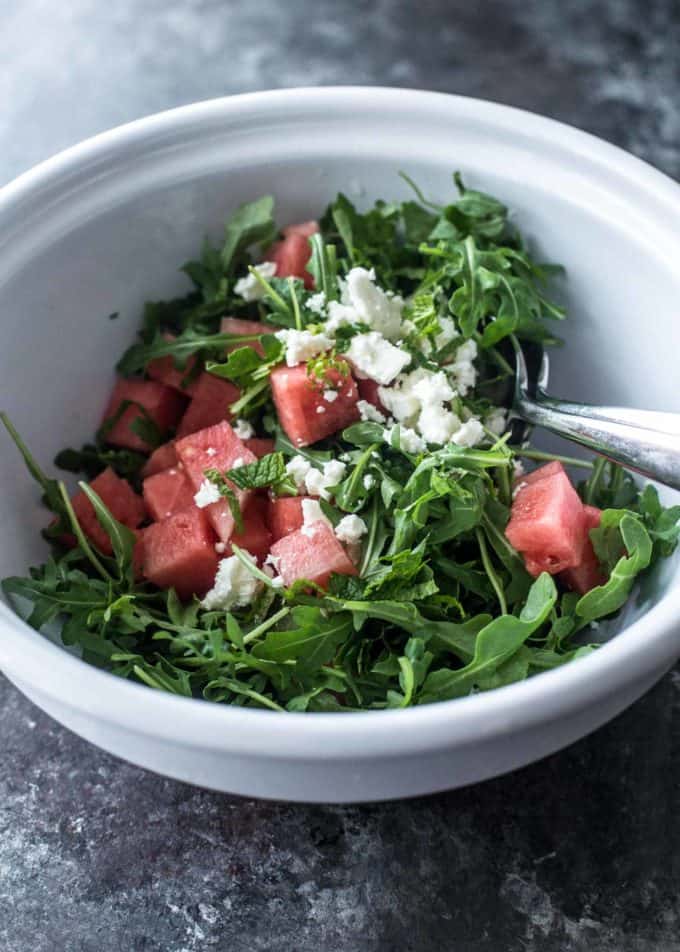 tossing watermelon, feta and arugula in a white bowl with two spoons