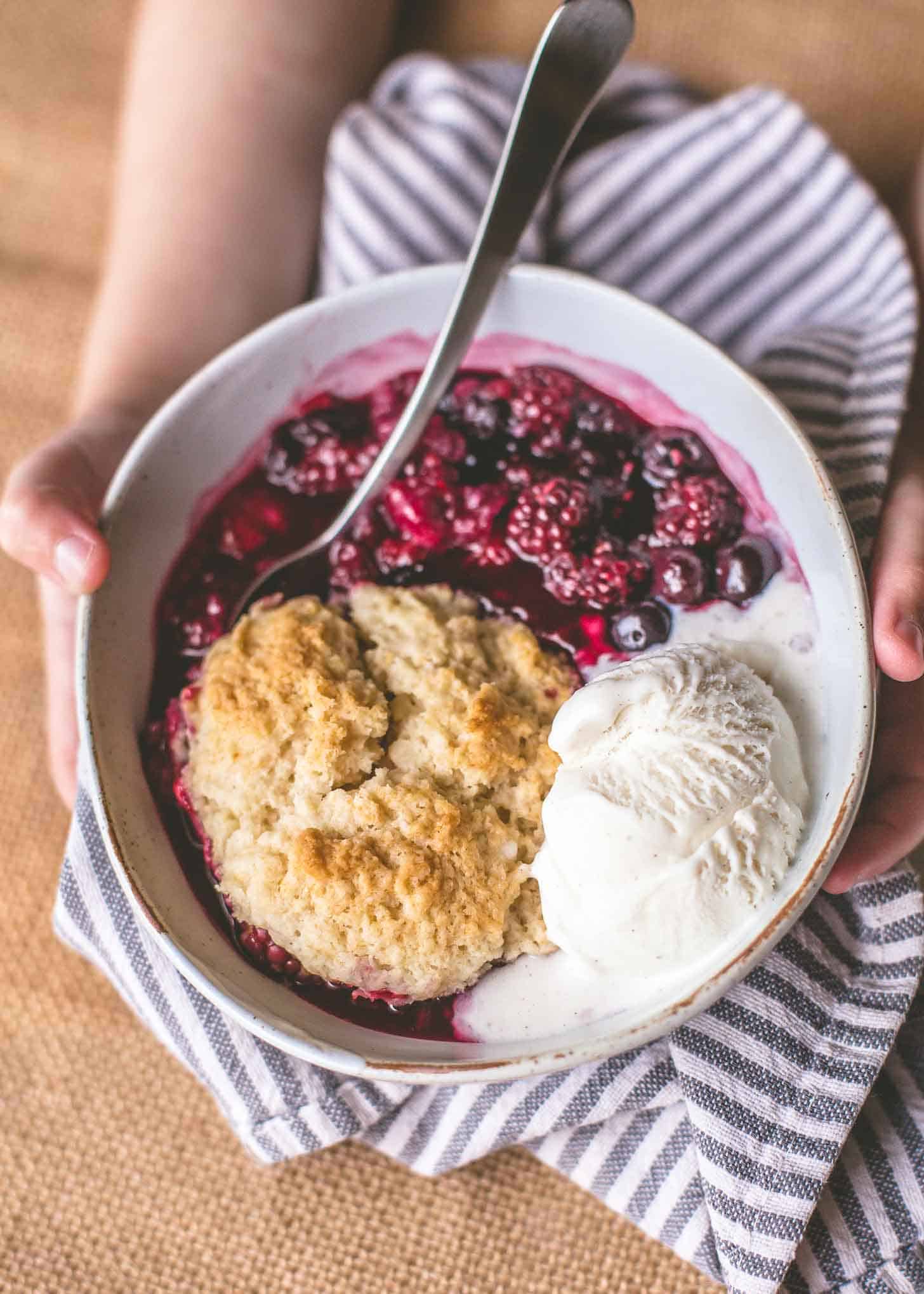 hands holding a bowl of berry cobbler with vanilla ice cream