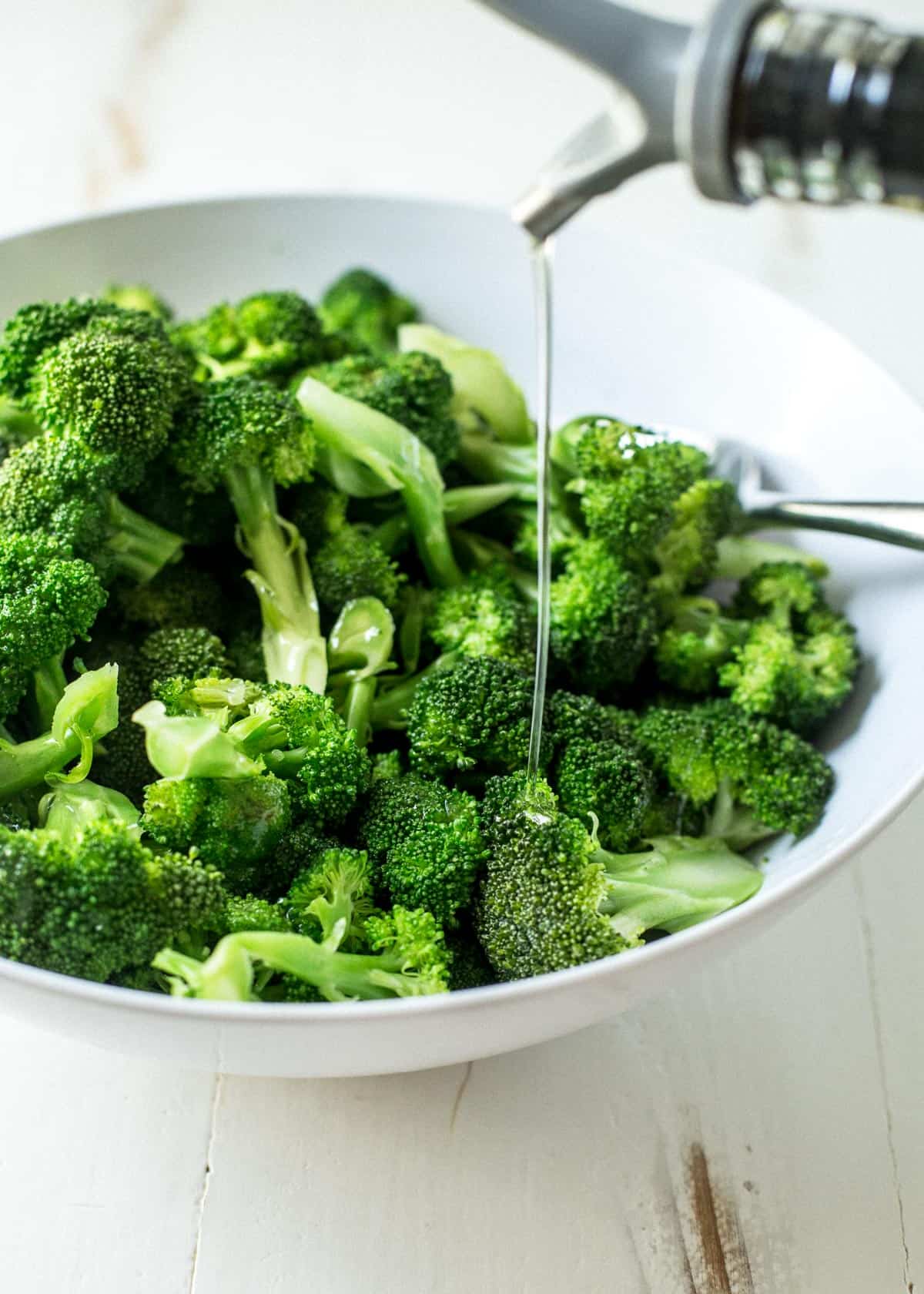 drizzling olive oil over raw broccoli in a white bowl