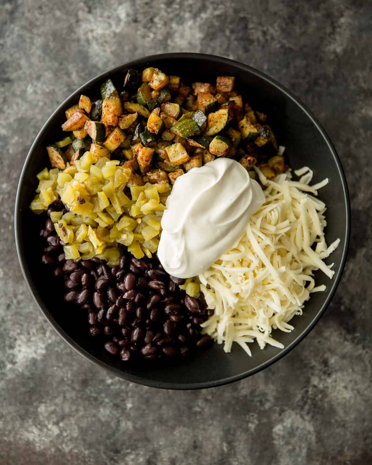 overhead image of black beans, zucchini, cheese, sour cream in a black bowl