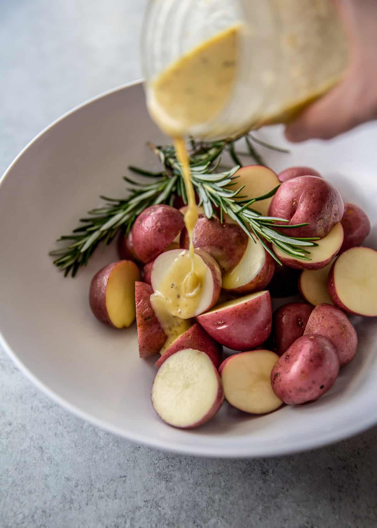 adding lemon dressing to potatoes and rosemary in a white bowl