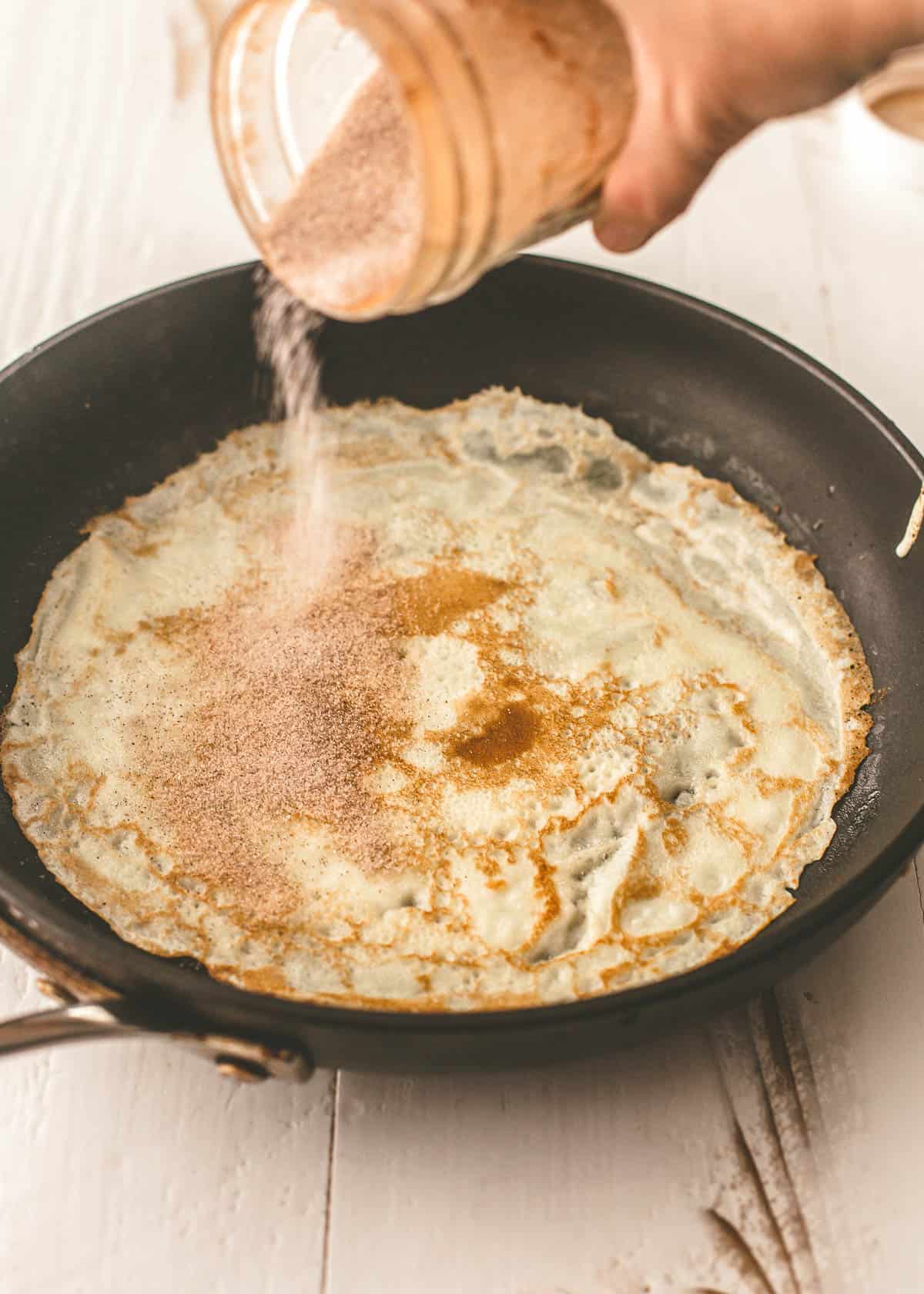 adding cinnamon sugar to crepe in a skillet