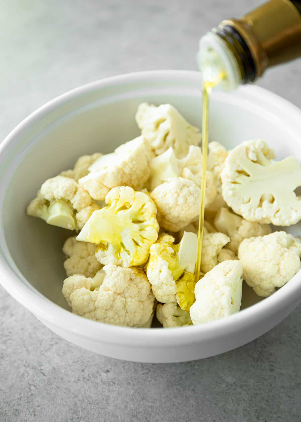 drizzling oil over raw cauliflower in a white bowl