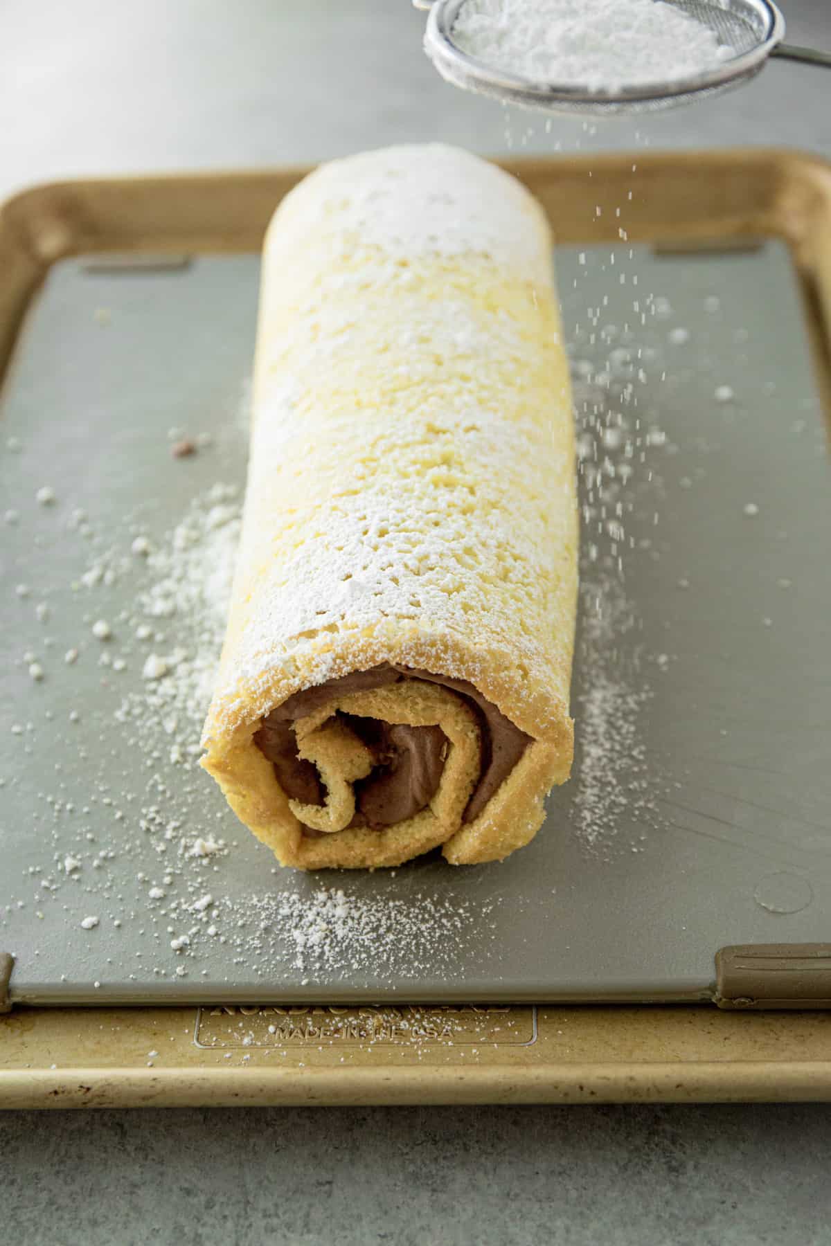 sprinkling powdered sugar over a cake roll on a sheet pan