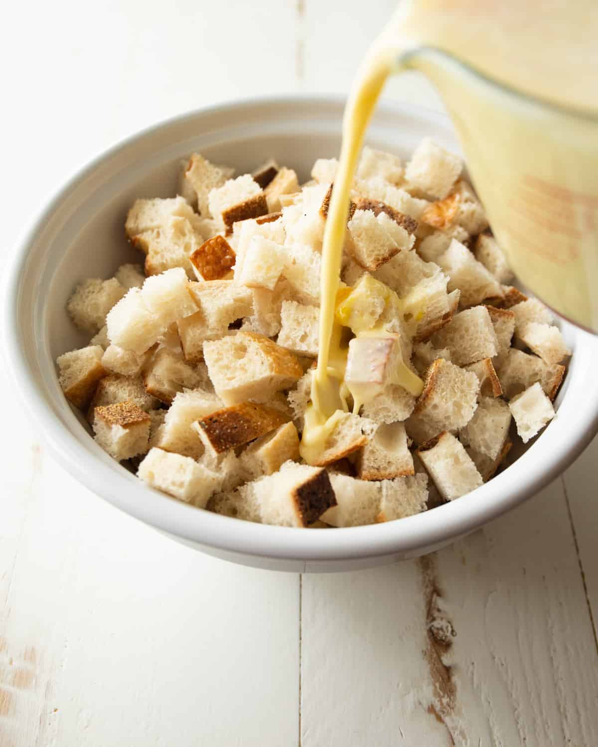 pouring egg mixture onto cubed bread in a white bowl