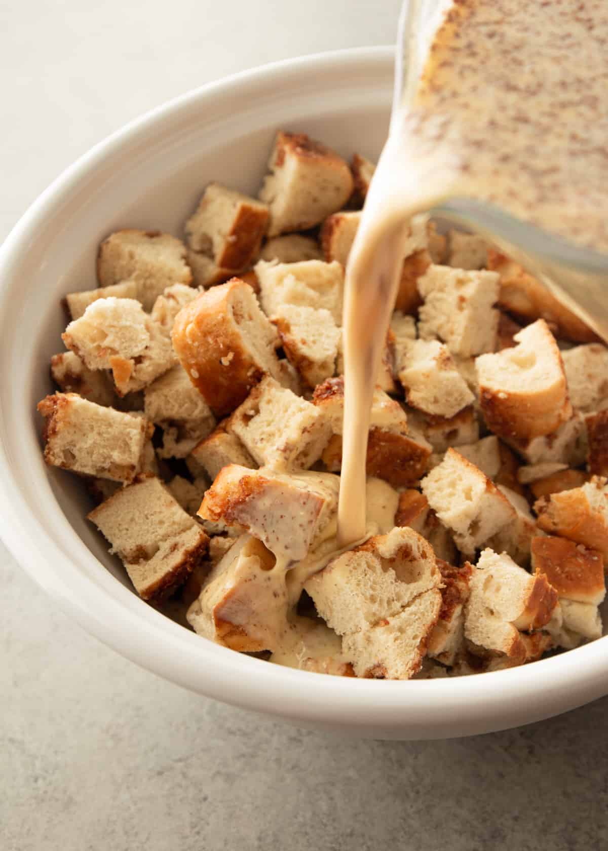 pouring egg mixture over cubed bagels in a white bowl