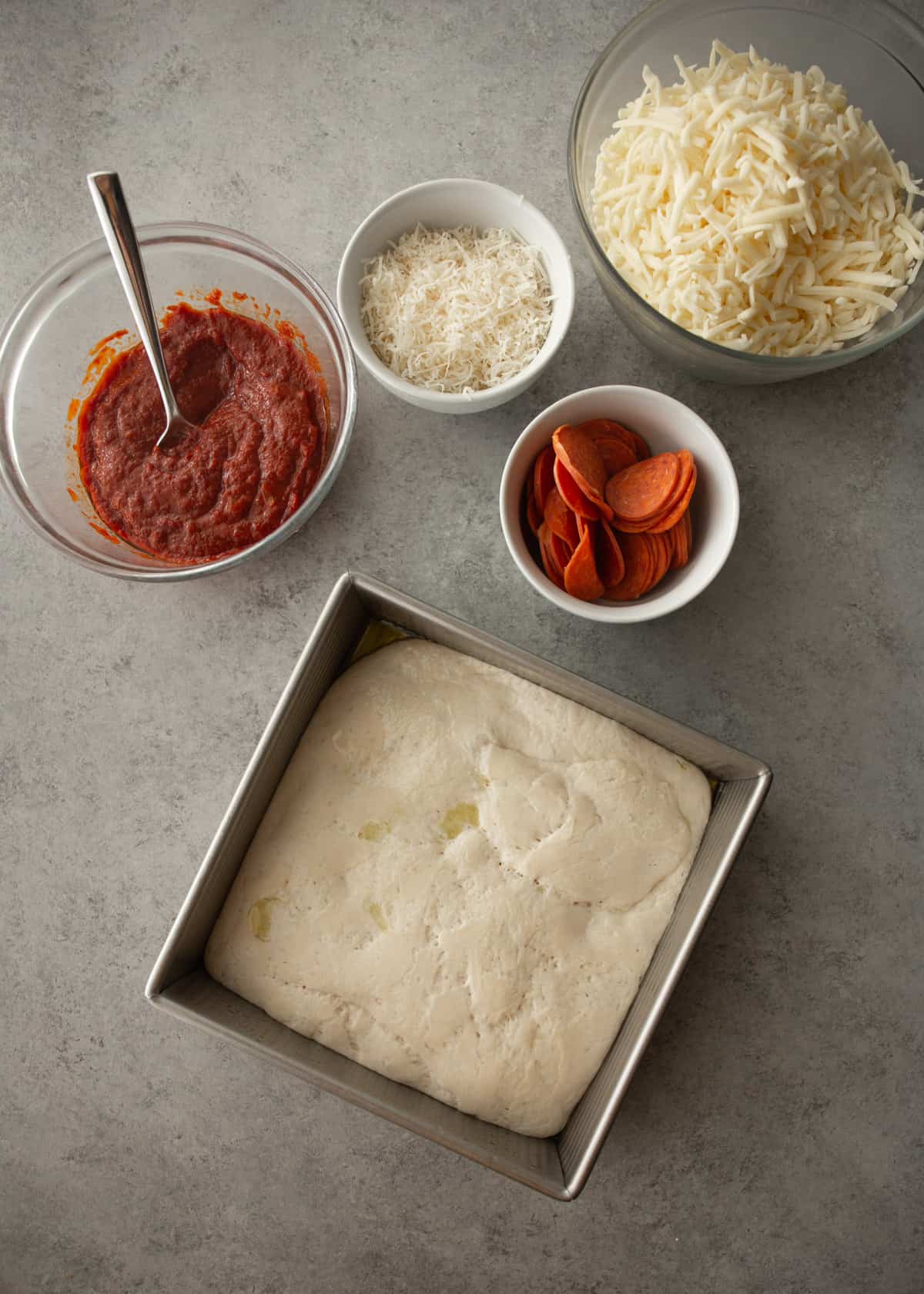 overhead image of dough, pepperoni, sauce and cheese on a grey countertop