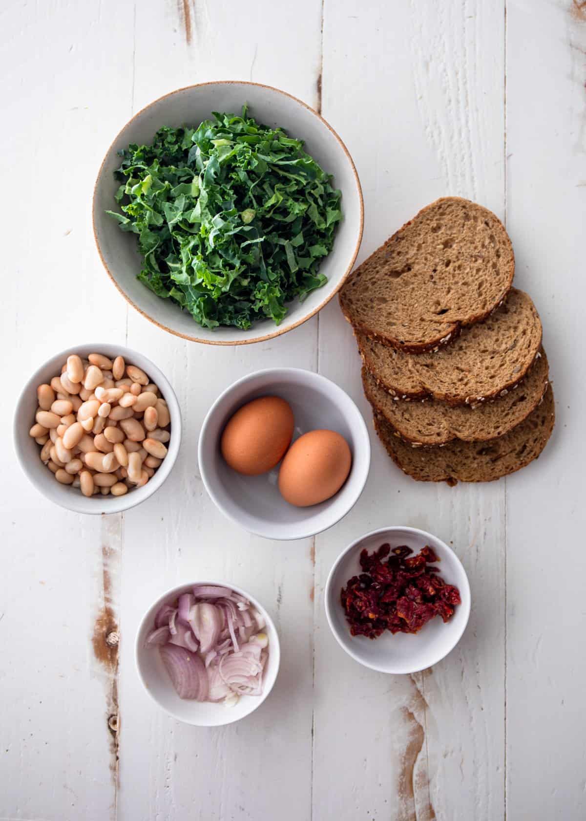 bread, beans, kale, shallots, eggs and peppers in small bowls on a white table