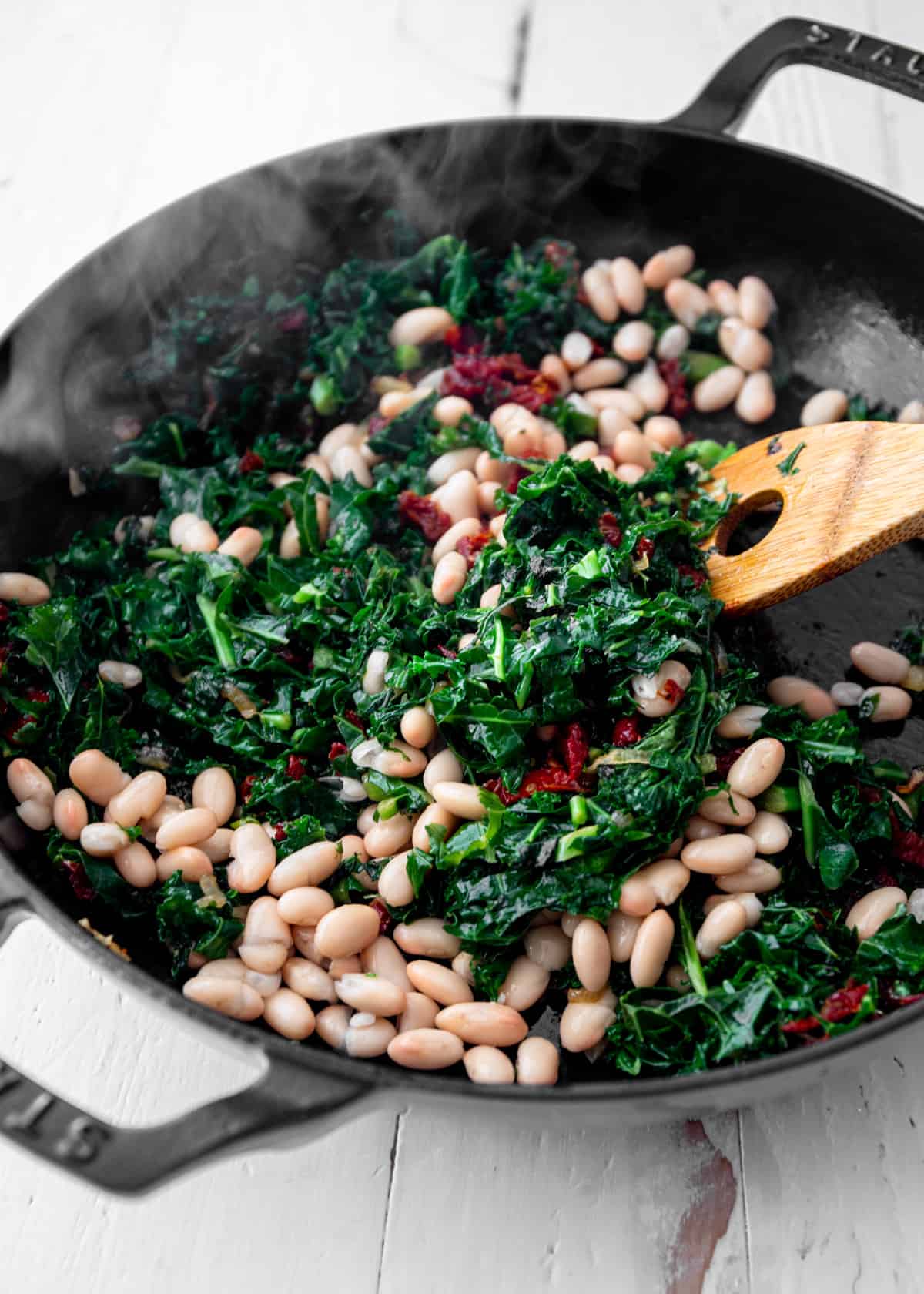 stirring kale, sun dried tomatoes and white beans in a cast iron skillet with a wooden spoon