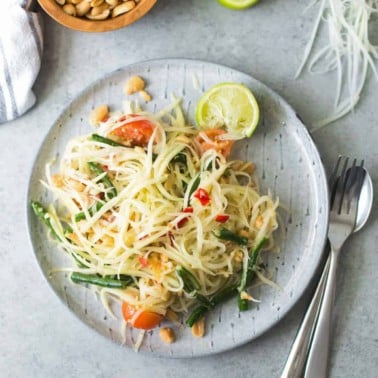 Thai green papaya salad on a blue plate with peanuts in a bowl on the side and lime wedges