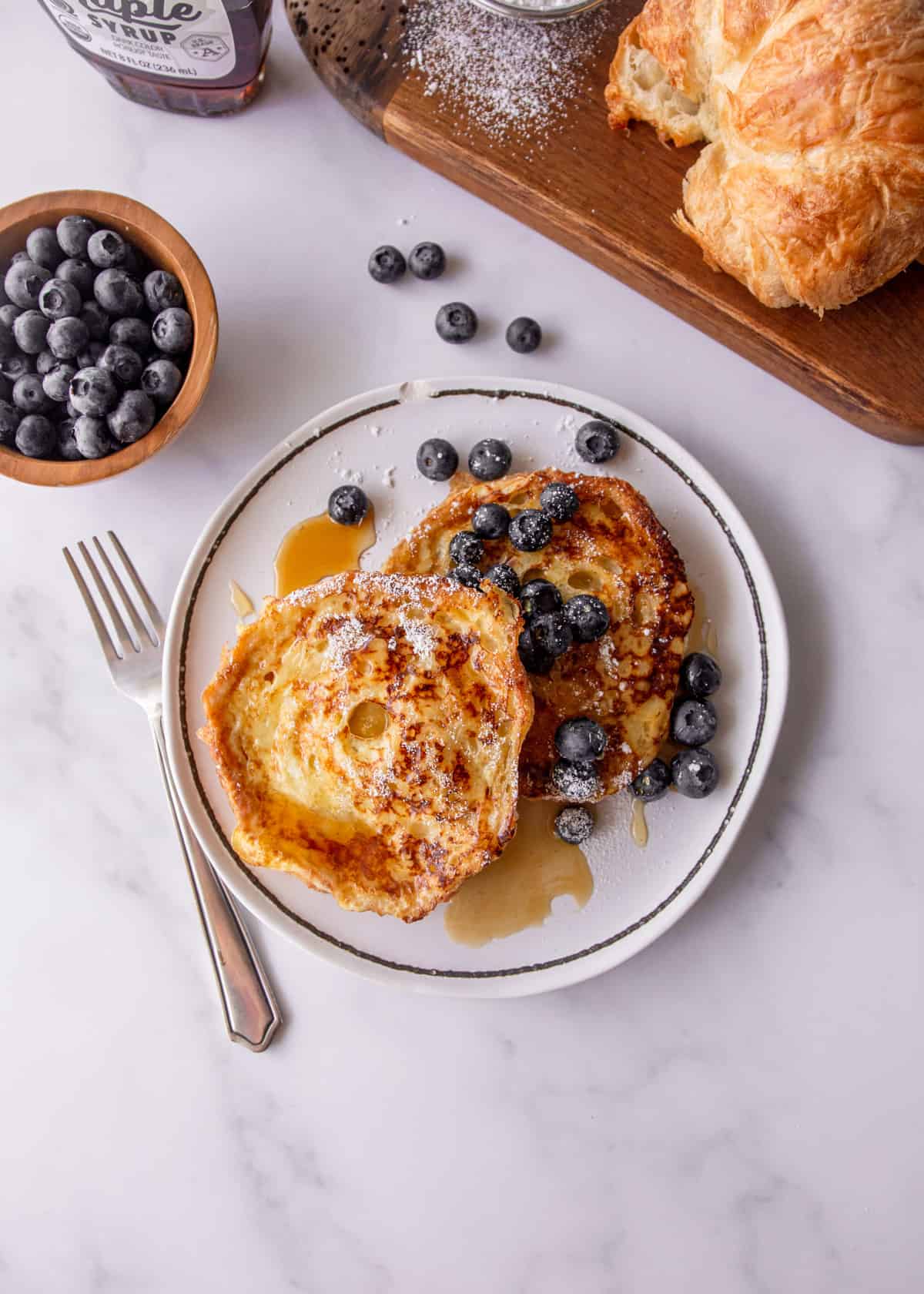 an overhead view of croissant french toast on a white plate with syrup and blueberries.