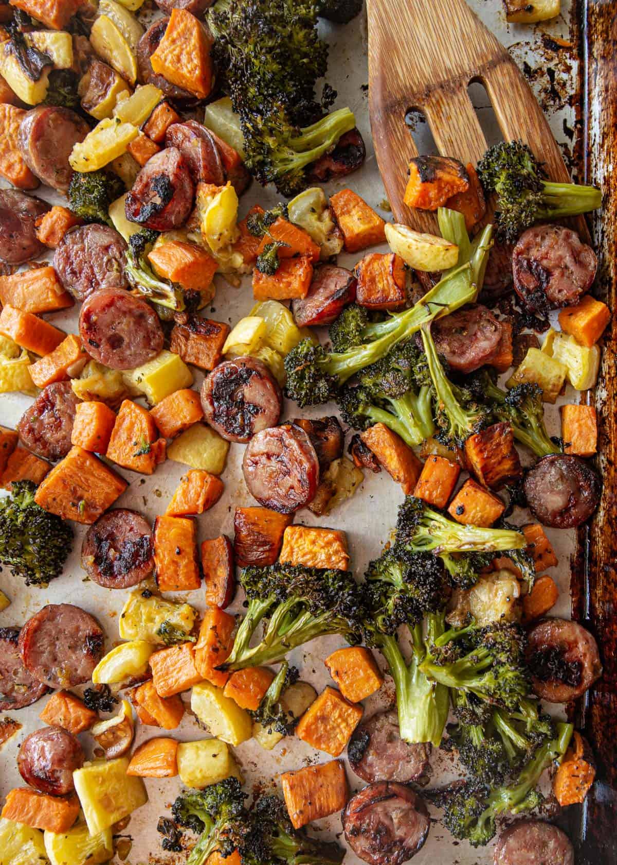 overhead image of a wooden spatula stirring roasted vegetables and meat on a sheet pan