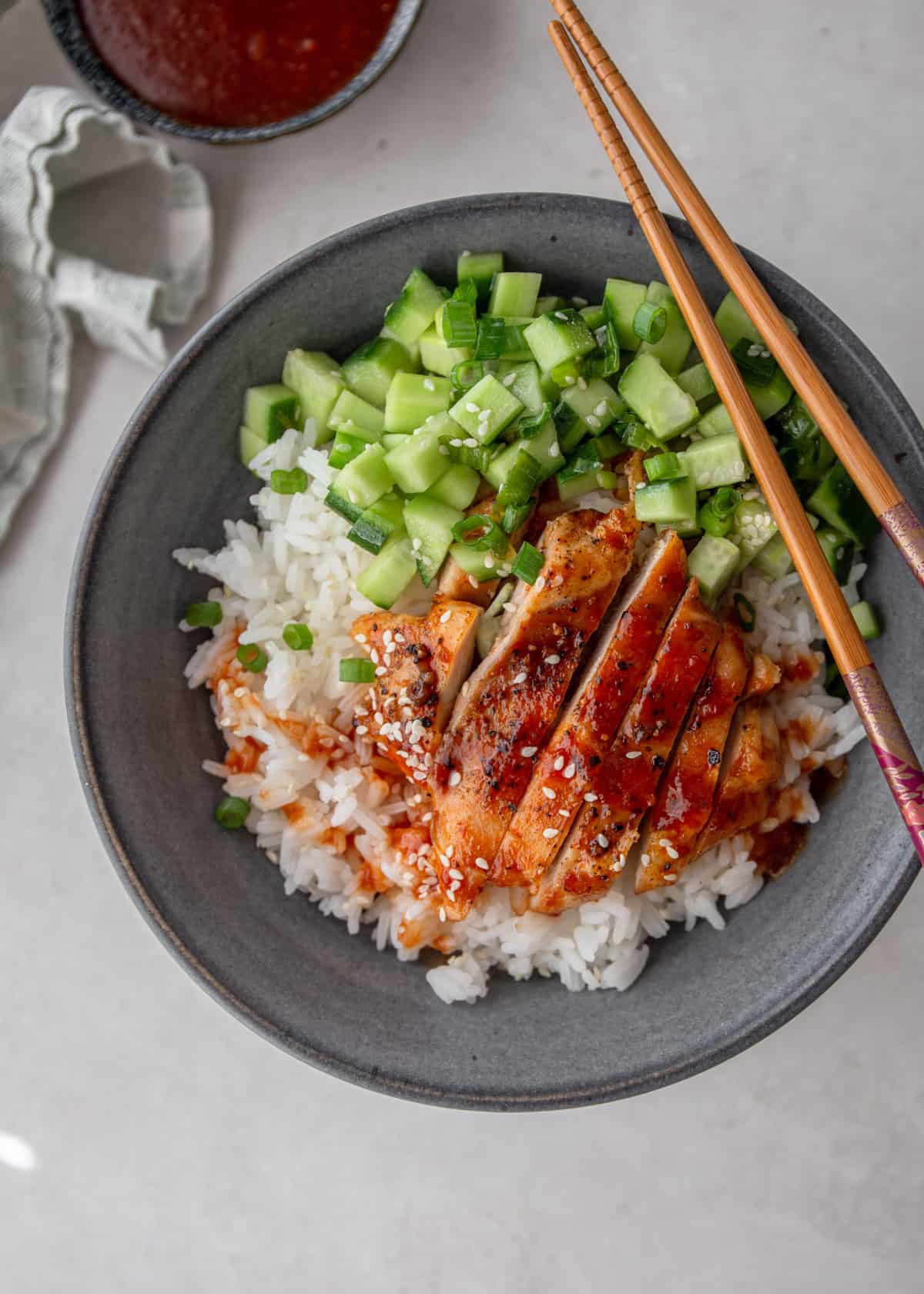 overhead image of glazed chicken slices, cucumbers and rice in a grey bowl with chopsticks.