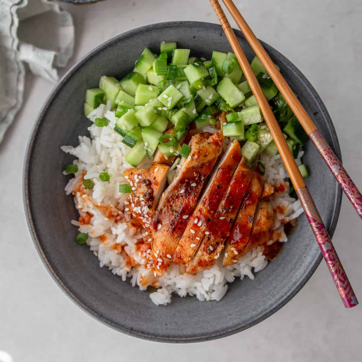 chicken slices, chopped cucumber and rice in a grey bowl with chopsticks