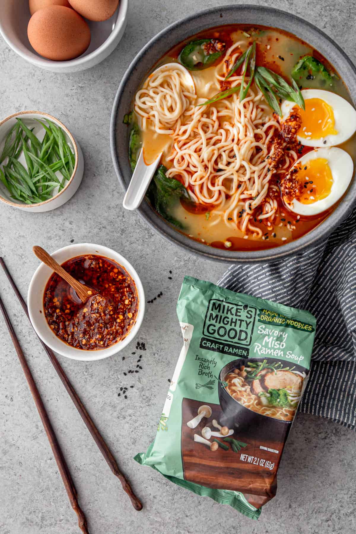 a bowl of ramen, next to a package of mike's mighty good ramen and a bowl of chili sauce