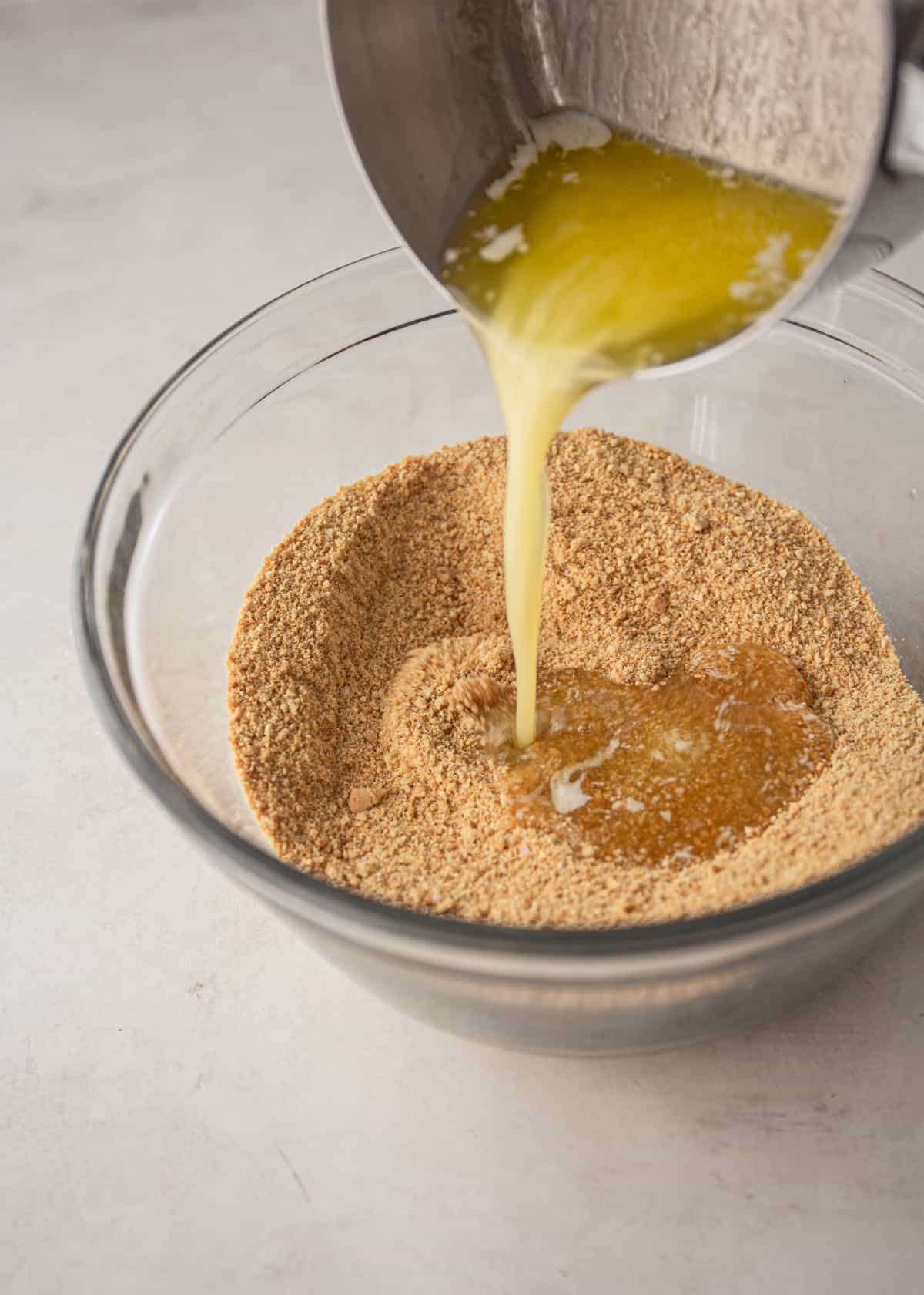 pouring melted butter into graham cracker crumbs in a clear glass bowl