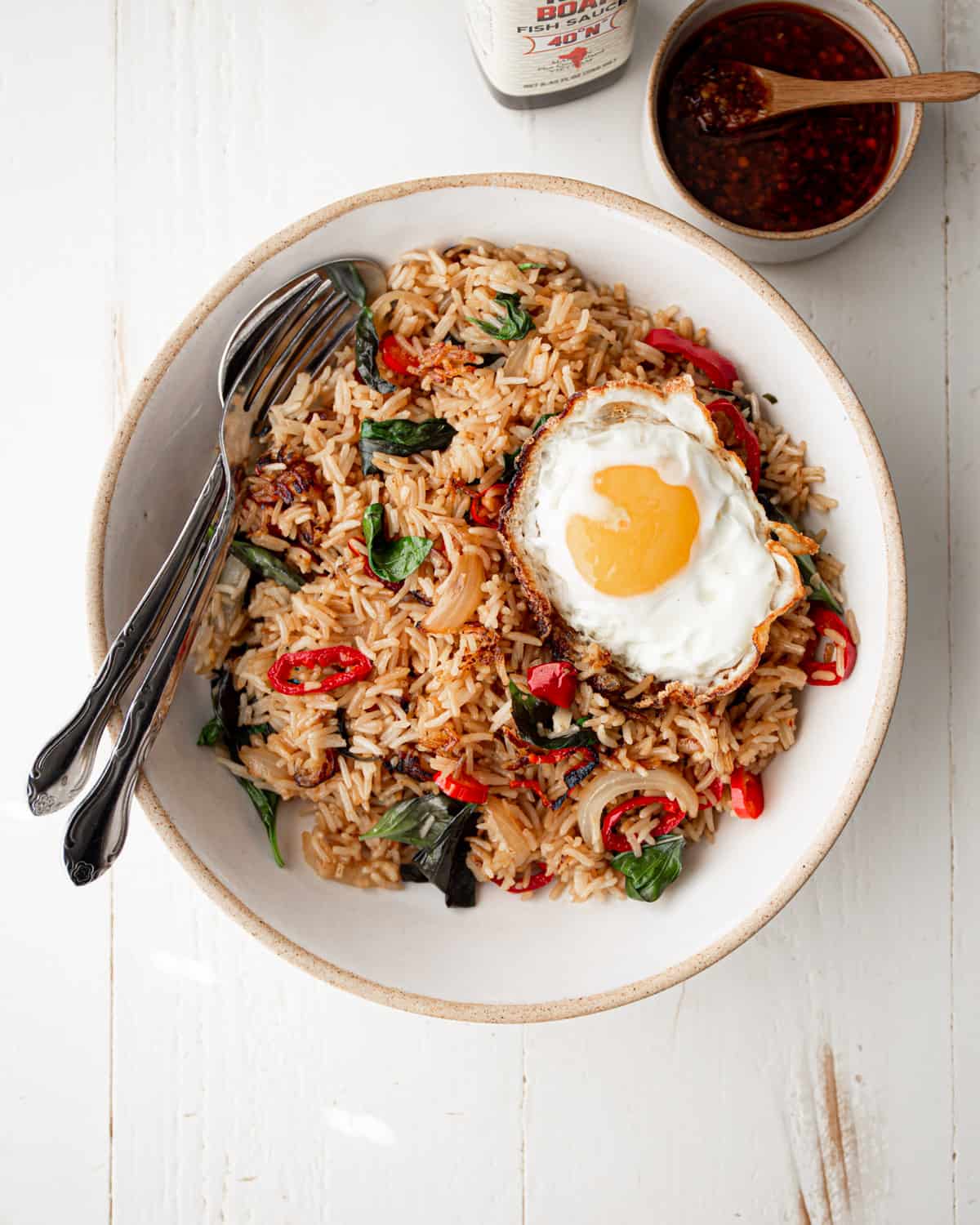 thai fried rice in a white bowl with a spoon and a fork, topped with a fried egg