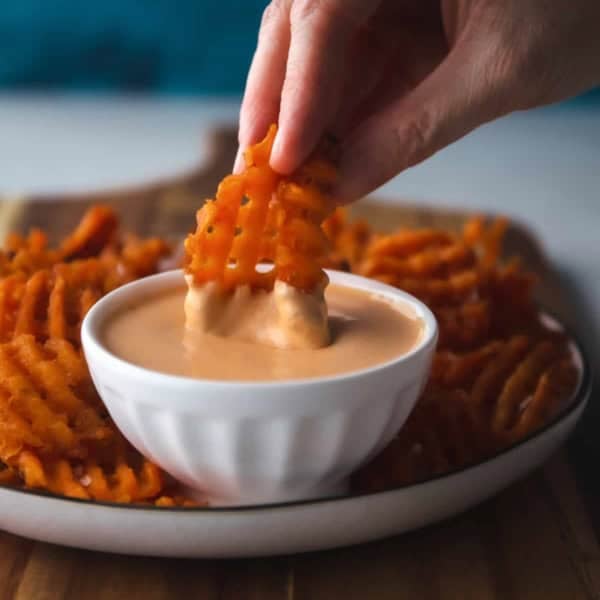 A sweet potato waffle fry dipped in a white bowl of mayo and Sriracha sauce