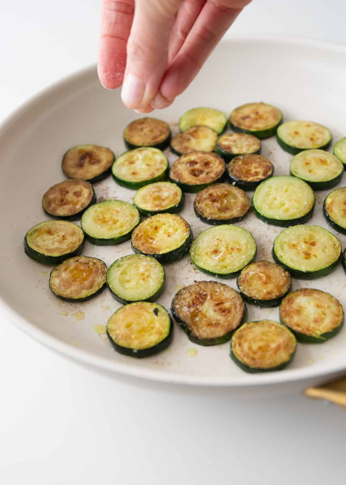 sprinkling salt on roasted zucchini slices in a round baking dish